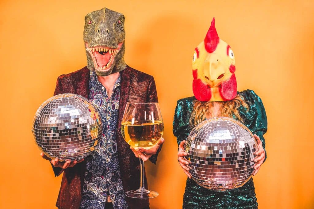 Crazy couple having fun celebrating new year eve party holding disco balls and glass of wine - Focus on t-rex and chicken mask