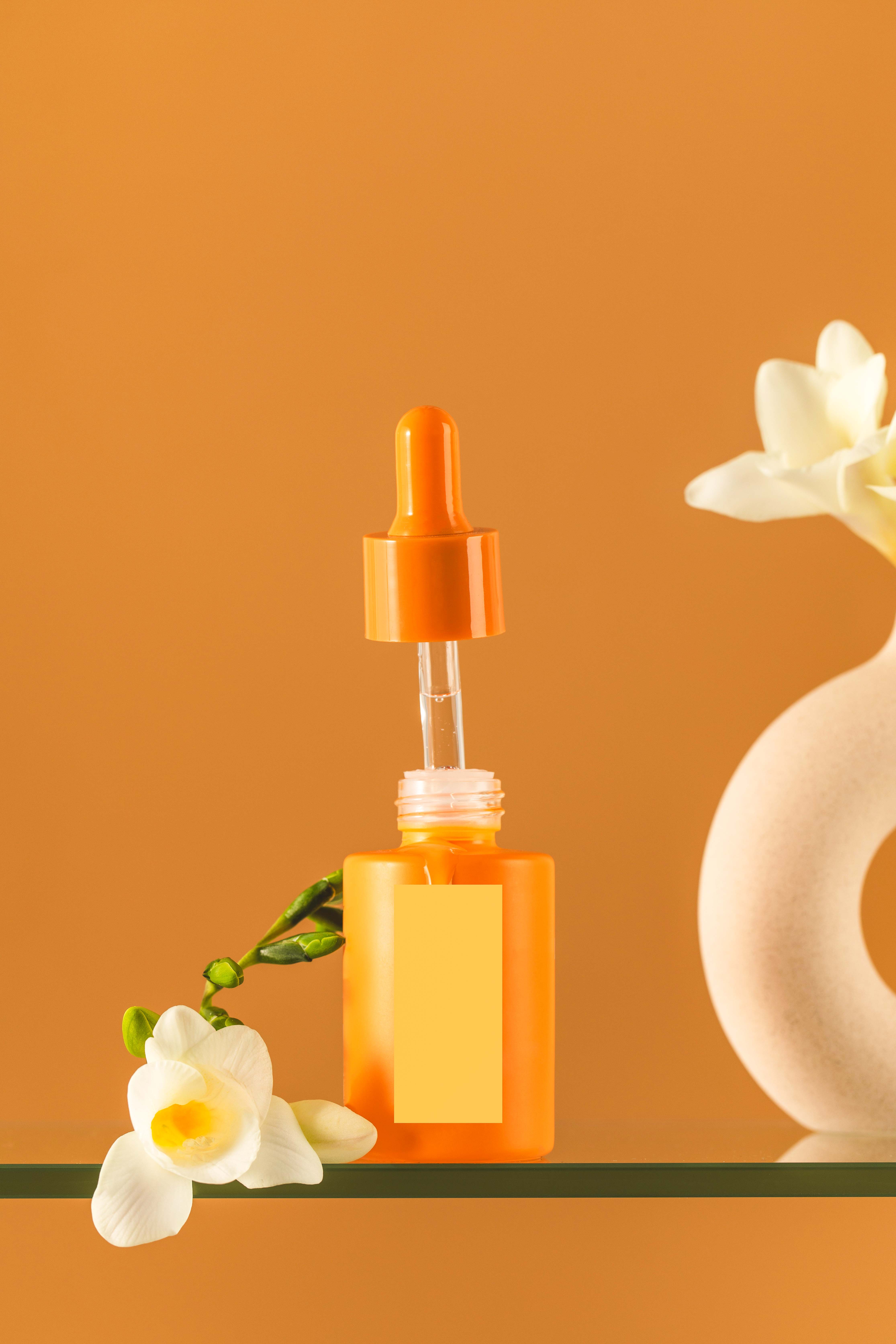 Aesthetic orange serum bottle next to minimalist vase with white flower on a glass shelf, trendy decor. Skin care